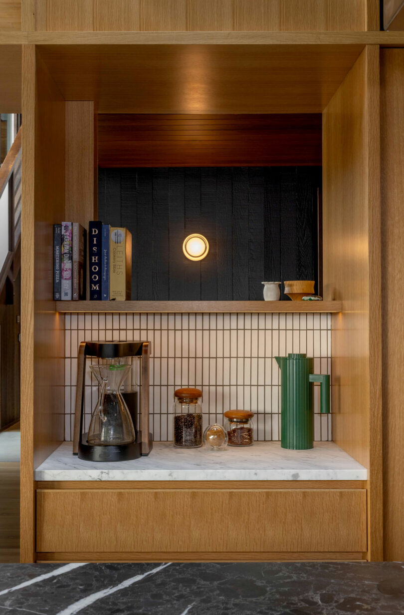 Modern kitchen shelf with books, a coffee maker, glass jars, and a green vase under a warm light, set against a textured black backdrop.