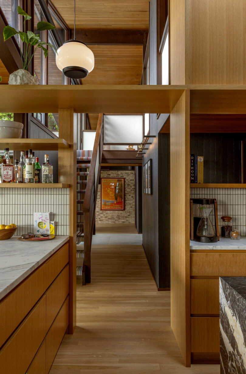 Modern kitchen with wooden cabinets and shelving, featuring a staircase in the background, white countertops, and small decorative items.