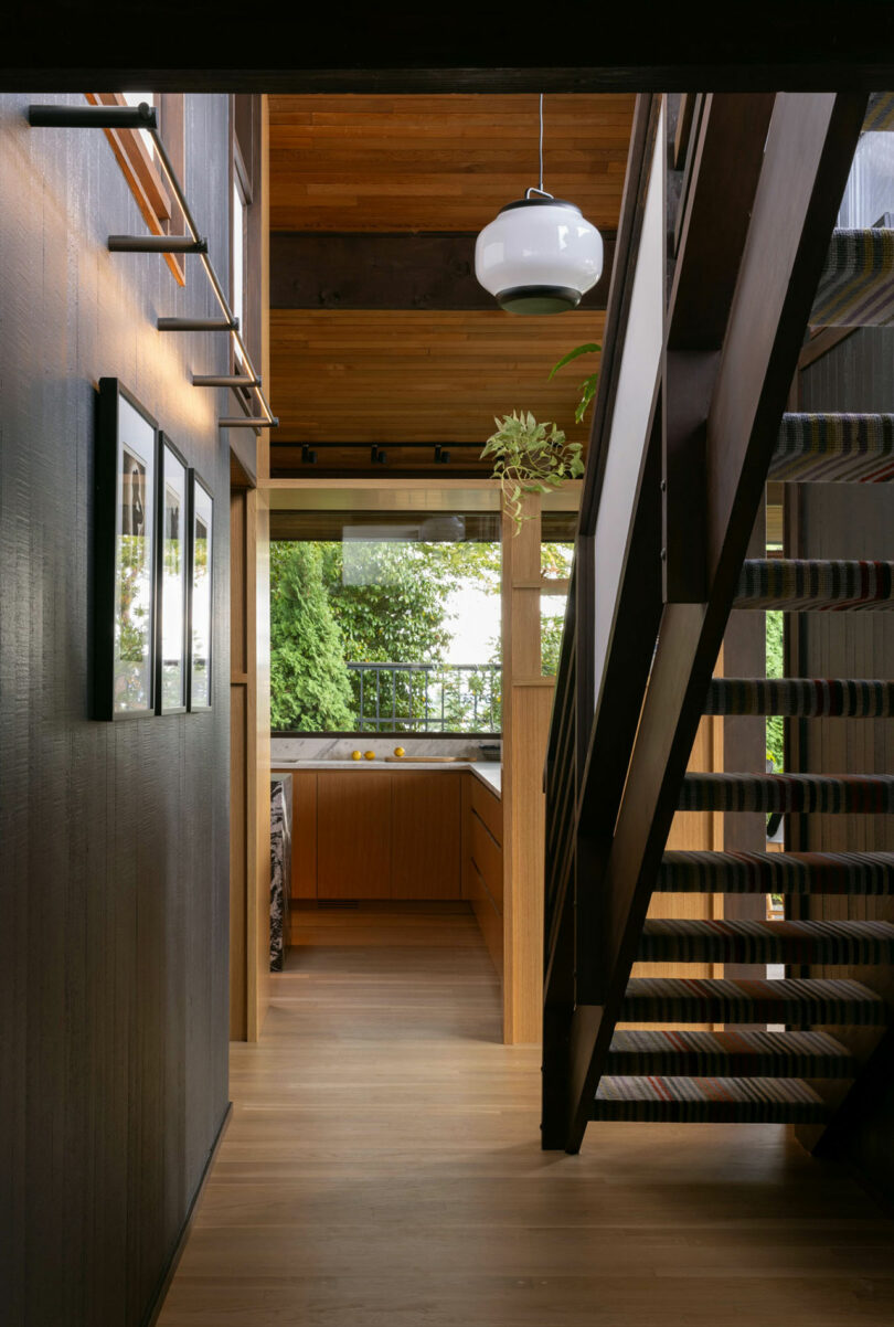 Modern interior hallway with wooden walls and floors, a staircase with black railings on the right, and framed pictures on the left.