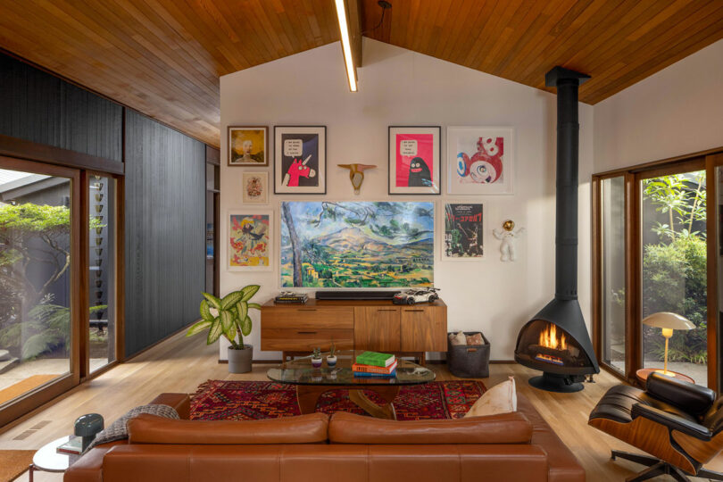 A cozy living room with a leather sofa, a freestanding fireplace, and walls decorated with various artworks, surrounded by large windows and lush greenery outside.