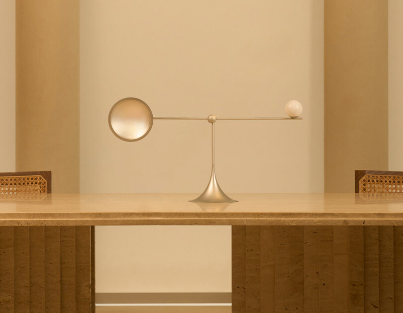 Um candeeiro de mesa minimalista Lumio Ovo com acabamento em ouro mate assenta sobre uma mesa de madeira, apresentando um design equilibrado com uma luz esférica e outra circular.