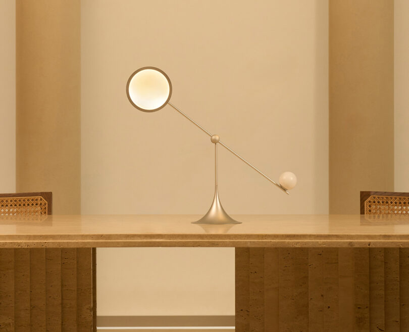 A modern Lumio Ovo table lamp with an elongated arm and two spherical ends, set on a wooden table against a beige wall angled upward to the left.