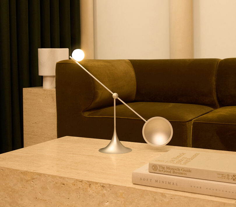 Um candeeiro de mesa Lumio Ovo com braço regulável ilumina um canto de uma sala com um sofá verde escuro e uma mesa de mármore com livros.