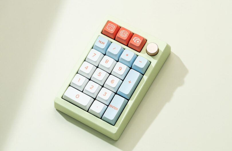 A minimalist green Meletrix ZoomPad Essential Edition keyboard with large, light blue and red buttons, displayed on a light beige background with a soft shadow.