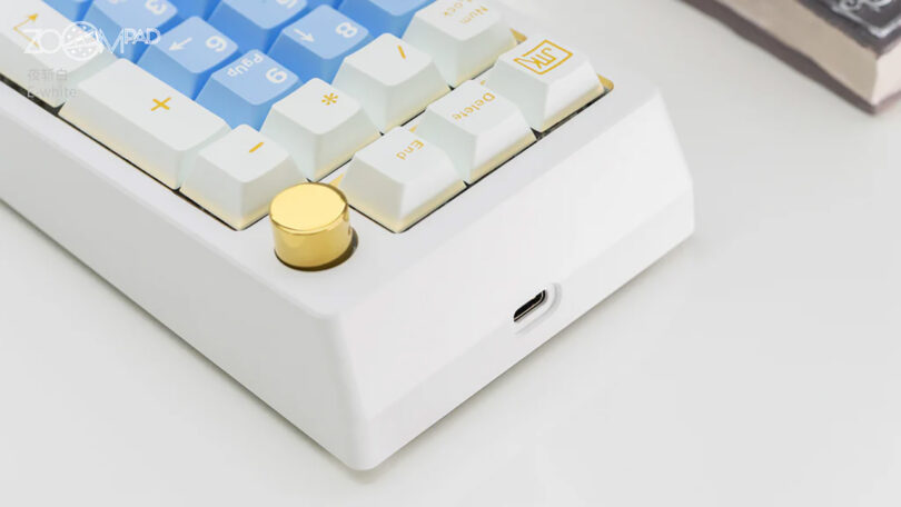 A close-up view of the Meletrix ZoomPad Essential Edition keyboard with white and blue keys, featuring a large gold-colored knob on the upper right side, and USB-C port