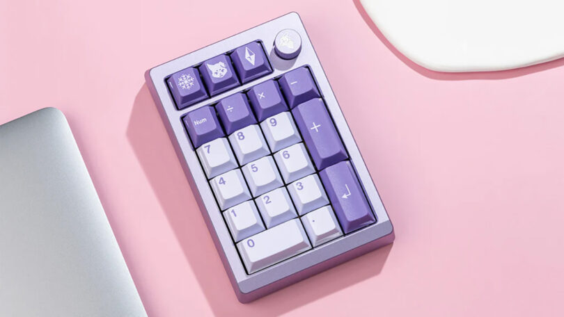 A Meletrix ZoomPad Essential Edition keyboard with purple and gray keys on a pink background, next to a laptop and white mouse pad.