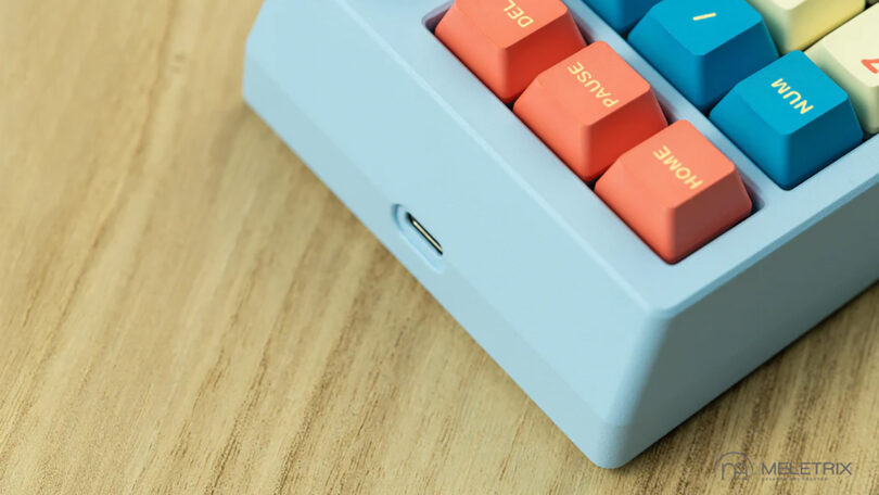 Close-up image of a blue Meletrix ZoomPad Essential Edition keyboard with colorful keys marked 'ctrl', 'del', 'home', and 'page up', displayed on a wooden surface, with USB-C port visible.