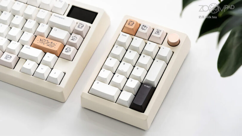 A minimalist desk setup featuring the Meletrix ZoomPad Essential Edition keyboard and a matching numpad with custom keycaps, set beside a houseplant on a white surface.