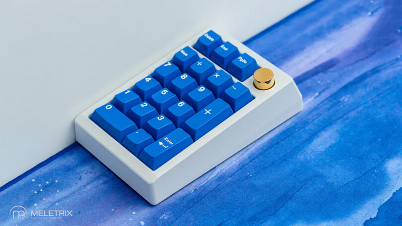 Meletrix ZoomPad Essential Edition keyboard and a matching numberpad with custom blue keycaps and gold dial, set across a blue watercolor painted surface.