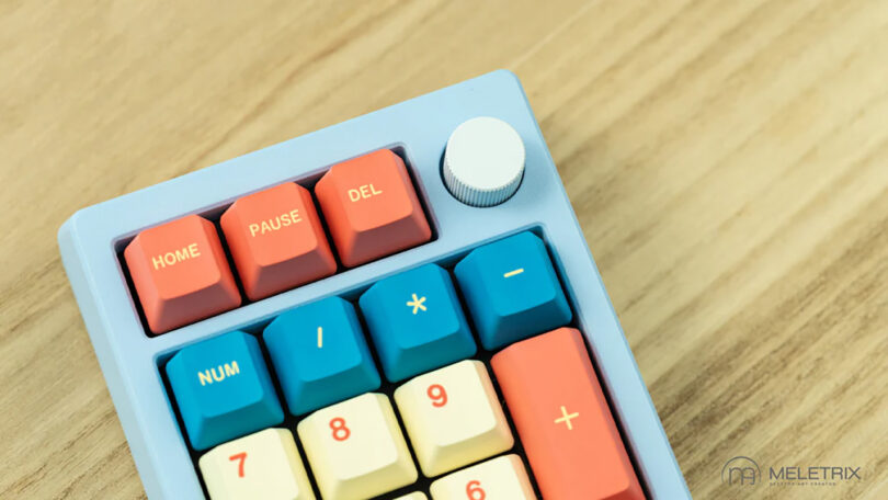 A colorful Meletrix ZoomPad Essential Edition keyboard with orange and teal keys, featuring symbols and function keys like home, pause, and del.