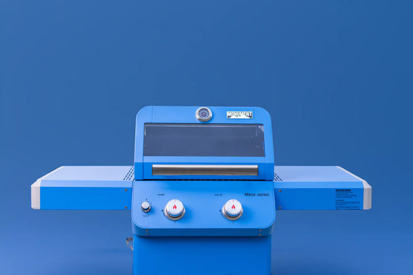 Blue Monument Grills Mesa Collection propane gas grill with both side tables extended set against a blue background.