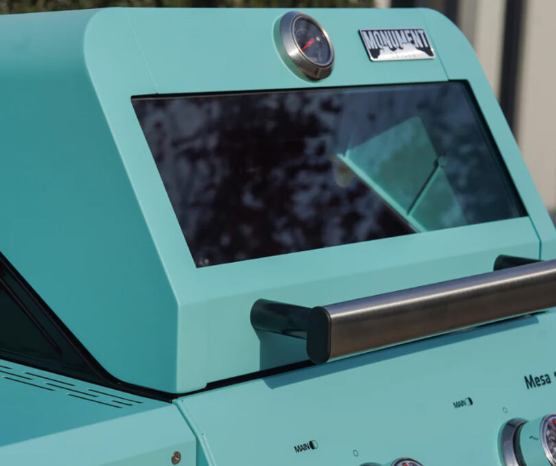 Close-up of a retro teal-colored Monument Grills Mesa Collection propane gas grill set outdoors with a reflection of trees visible across its glass window.
