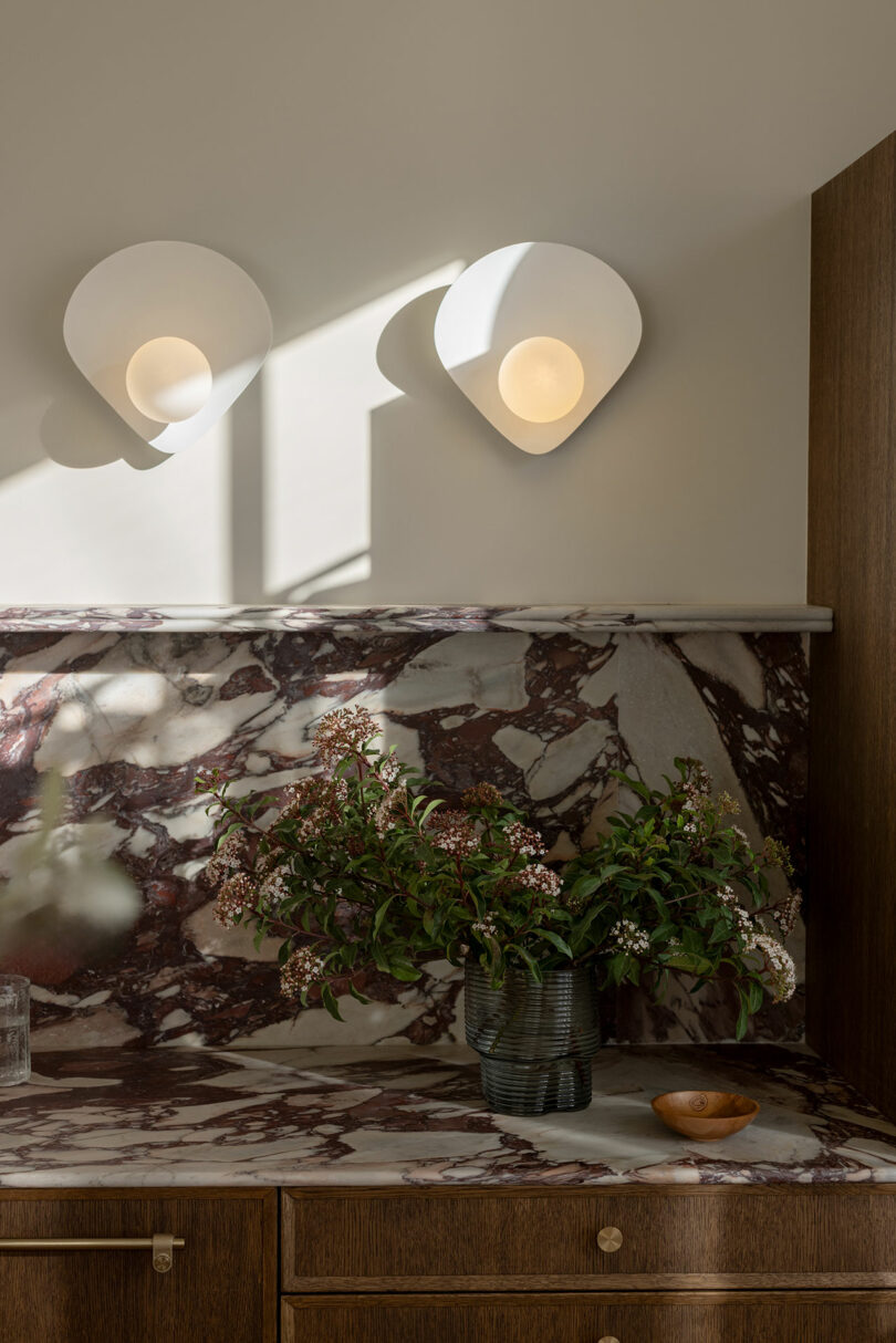 A stylish interior featuring two wall-mounted lights with a floral arrangement on a marbled countertop underneath.
