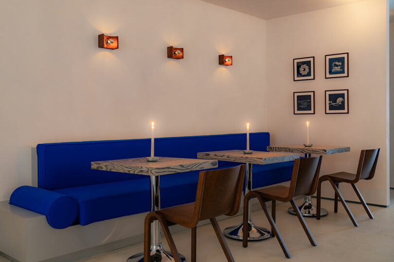 Modern dining area with blue upholstered bench, wooden chairs, marble tables, candles, wall lamps, and blue-framed artwork.