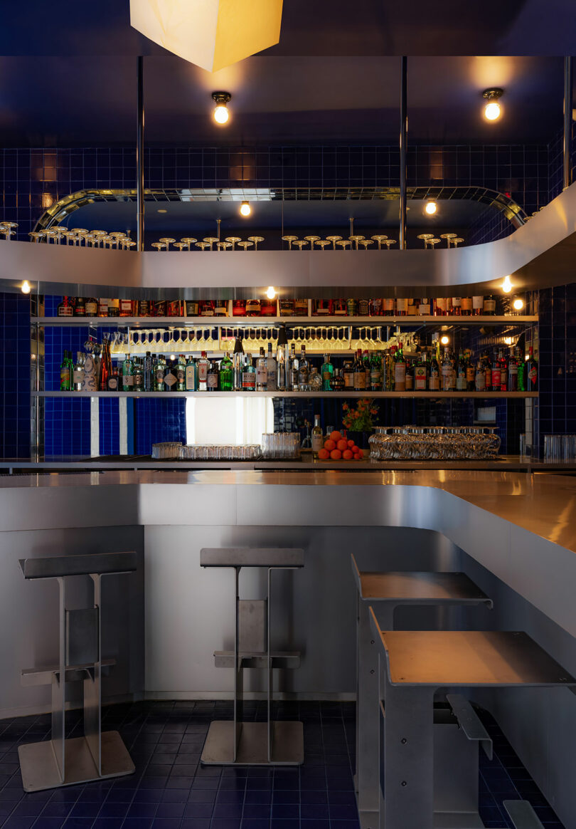 Modern bar interior with blue tile walls, backlit shelves with alcohol bottles, and stools in front of a white counter.