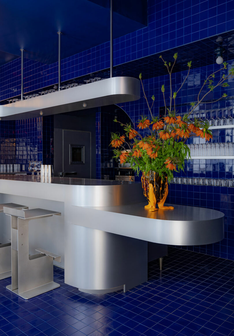 Modern bar interior with blue tile walls and silver counter, decorated with orange flowers in a yellow vase.