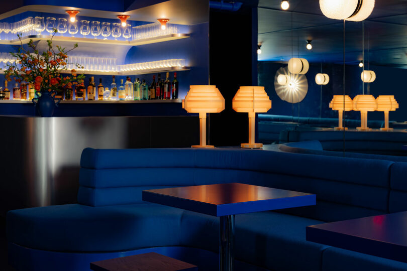 A stylish bar interior with dim lighting, featuring an illuminated counter, blue booths, and orange table lamps.