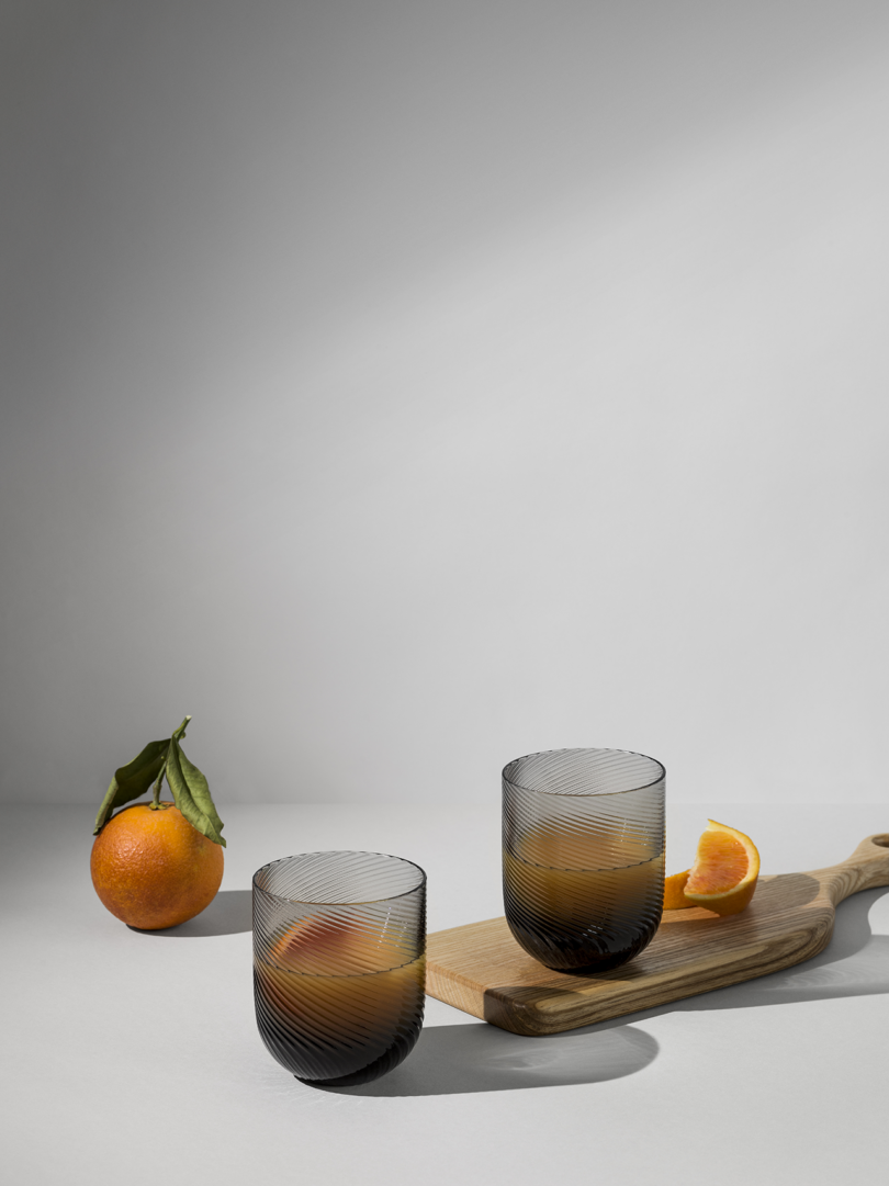 Two ribbed glasses on a wooden serving board with a whole and a halved orange.
