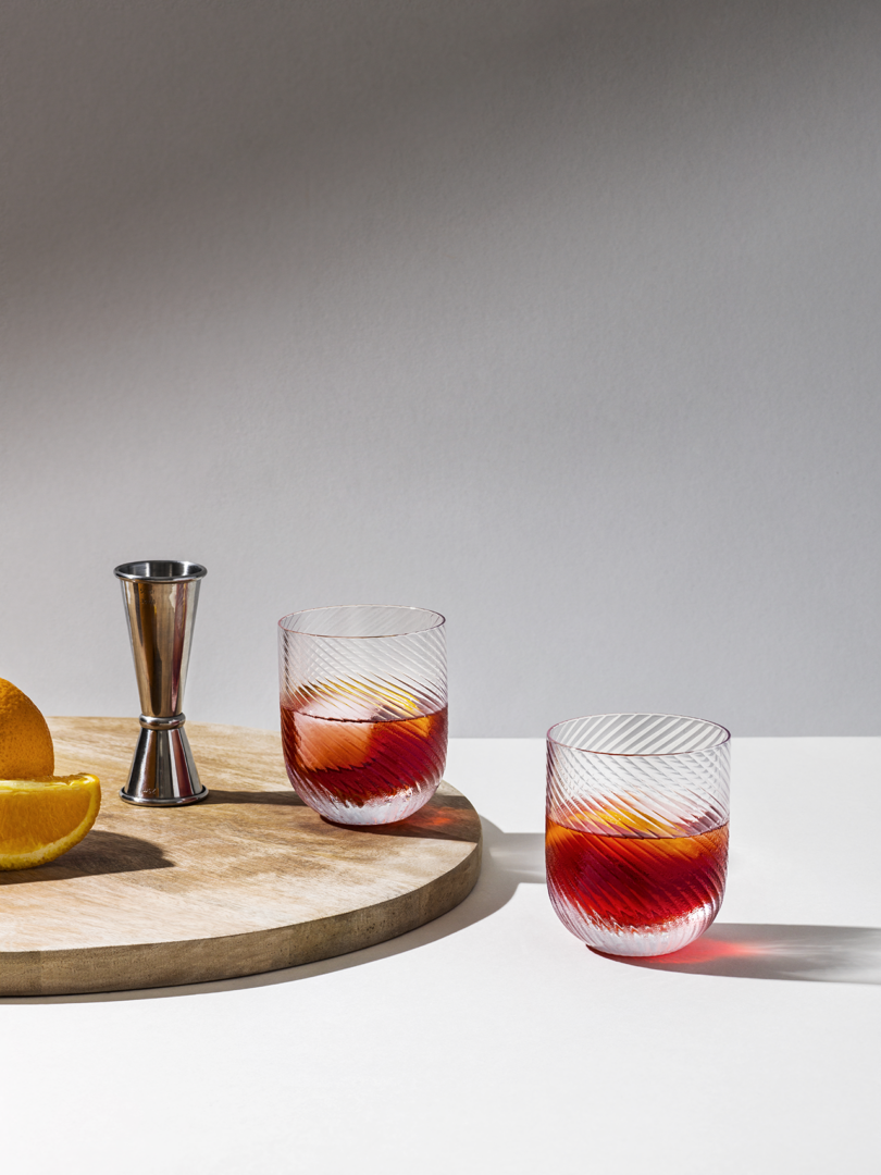 Two glasses with red liquid next to a cocktail jigger, on a surface with orange slices.