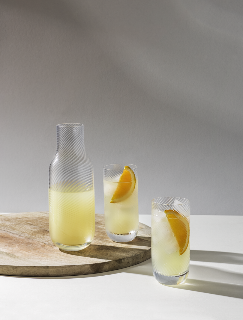 A carafe and two glasses of a citrus-infused beverage, garnished with orange slices, on a sunlit table.