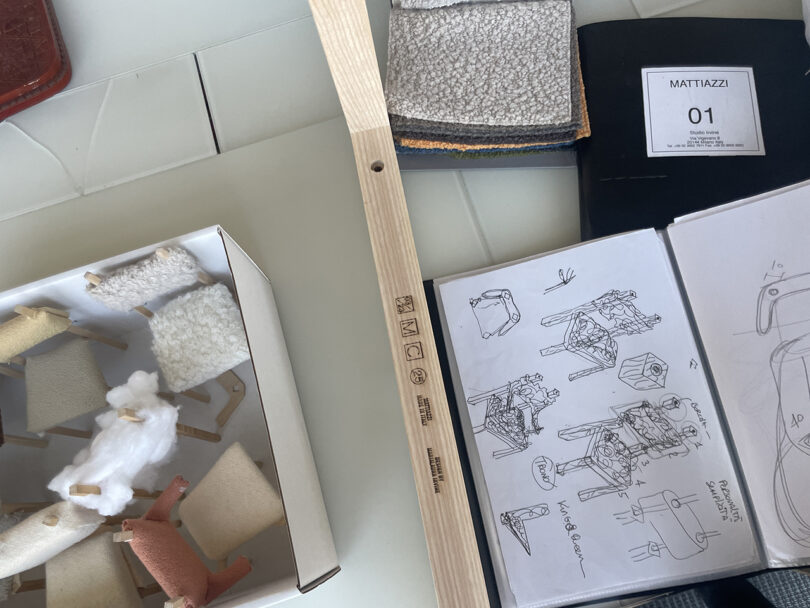 Aerial view of a design workspace with fabric samples, sketchbooks, and a wooden ruler on a white table.
