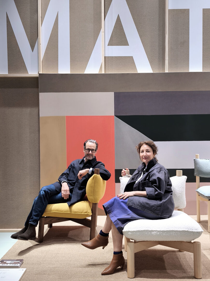Two people seated in a modern furniture showroom with large text on the wall behind them.
