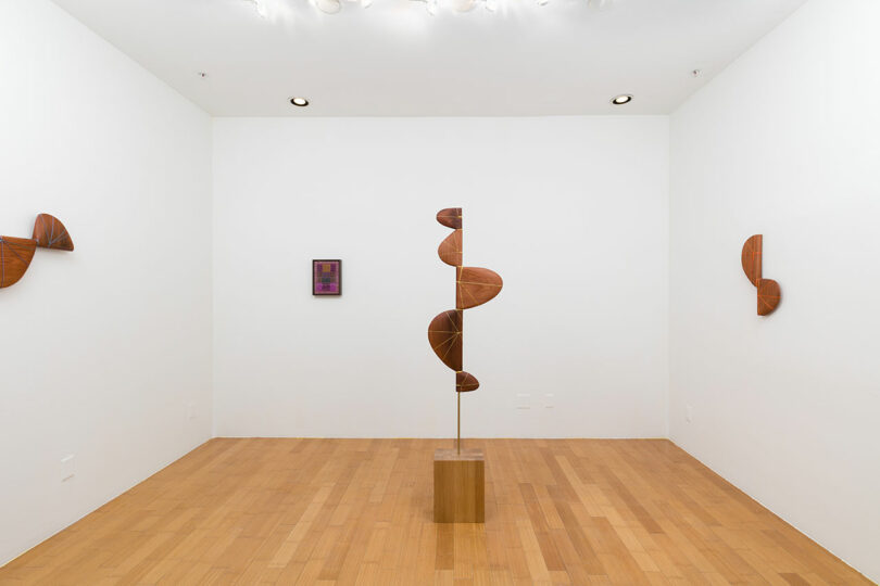 Art gallery room with a spiral wooden sculpture on a pedestal in the center and four abstract wooden wall sculptures.