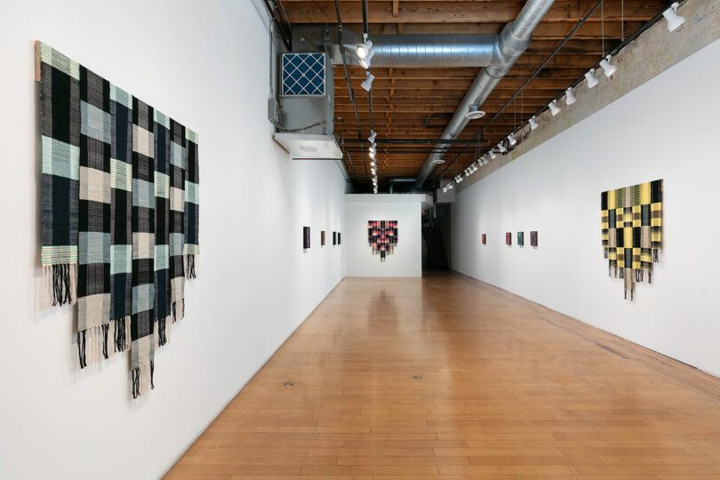 Interior of an art gallery with a series of textile artworks displayed on white walls alongside a long corridor with wooden flooring.