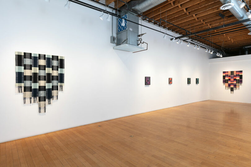 Art gallery interior with wooden floors and white walls displaying various artworks, including a large woven textile and several framed paintings.
