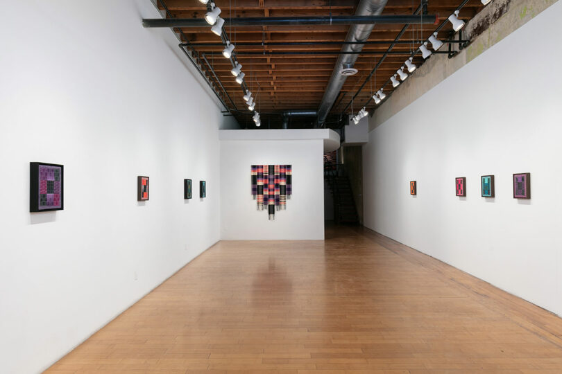 Interior of a modern art gallery with white walls displaying colorful abstract paintings and a wooden floor leading to a staircase at the back.