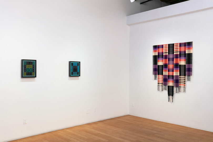 Interior of an art gallery showing a white wall with three artworks: two framed pieces on the left and a colorful textile artwork on the right.