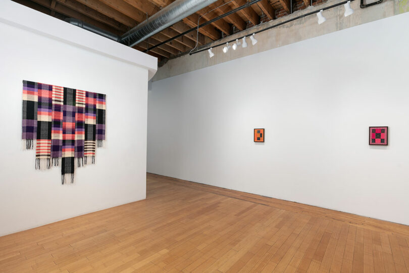 Modern art gallery interior with colorful woven textile on a white wall, and two framed artworks hanging on adjacent walls, in a room with wooden flooring.
