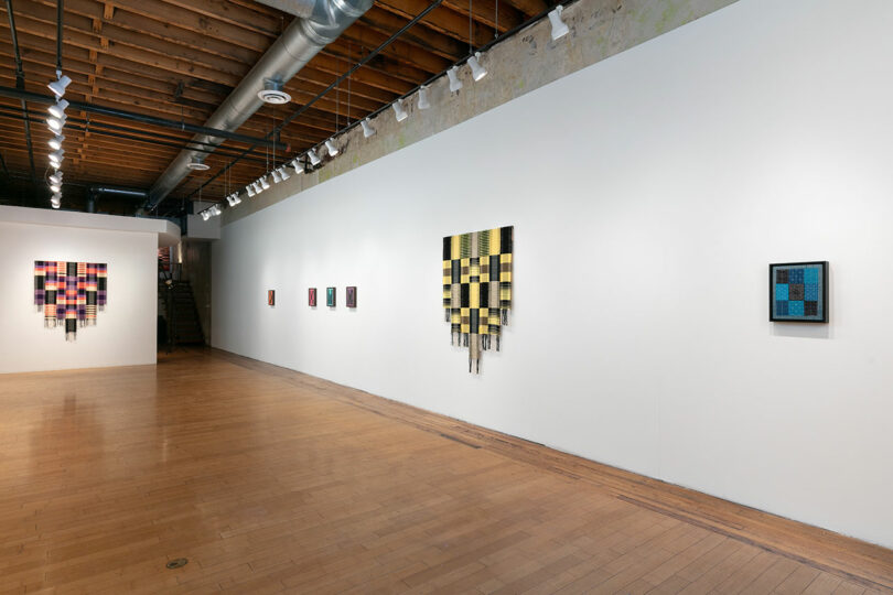 Modern art gallery interior showcasing colorful abstract paintings on a white wall, with exposed wooden beams and ductwork overhead.