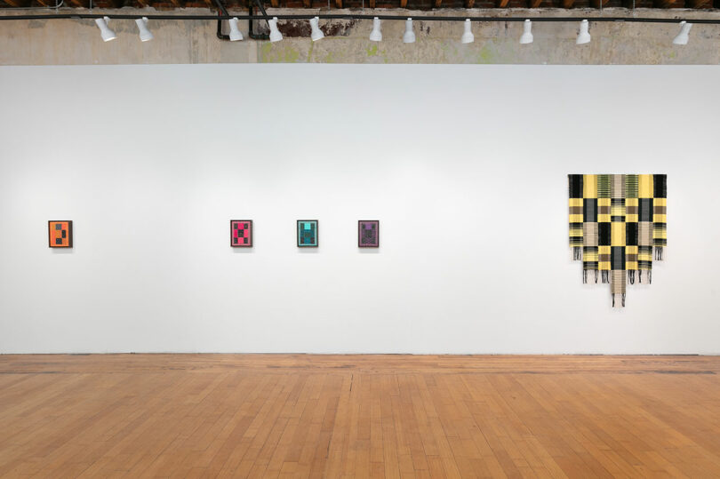 Art gallery interior with five colorful abstract paintings hung on a white wall, the largest featuring a black and yellow grid pattern on the right.