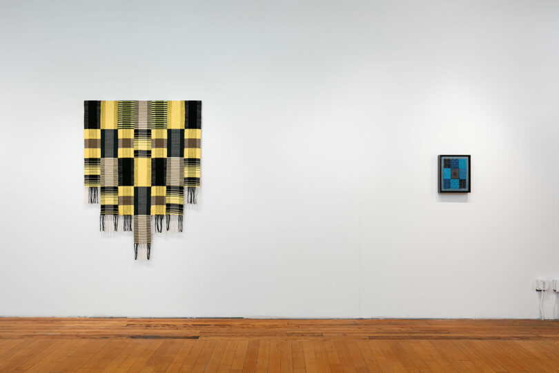 Two pieces of modern art on a white gallery wall; one large striped textile and a smaller framed blue piece to the right.