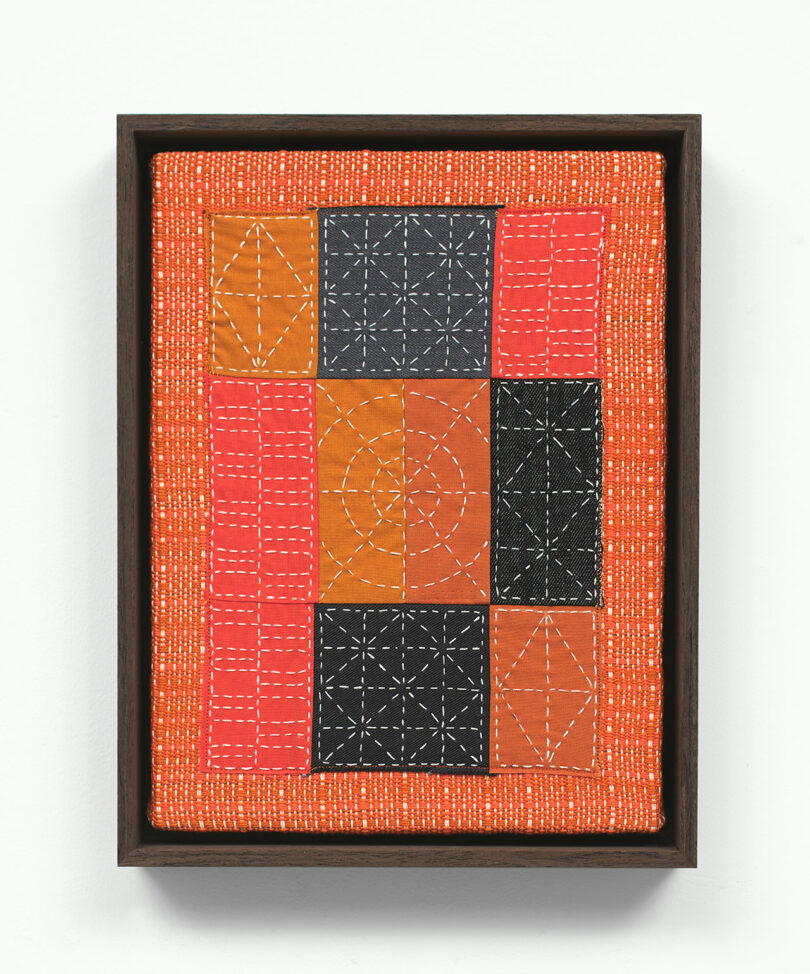 Framed textile artwork featuring a geometric patchwork of orange and black squares with intricate white stitching patterns.