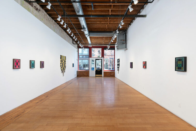 Interior of a bright art gallery with colorful abstract paintings on white walls, wooden floors, and exposed ceiling beams.