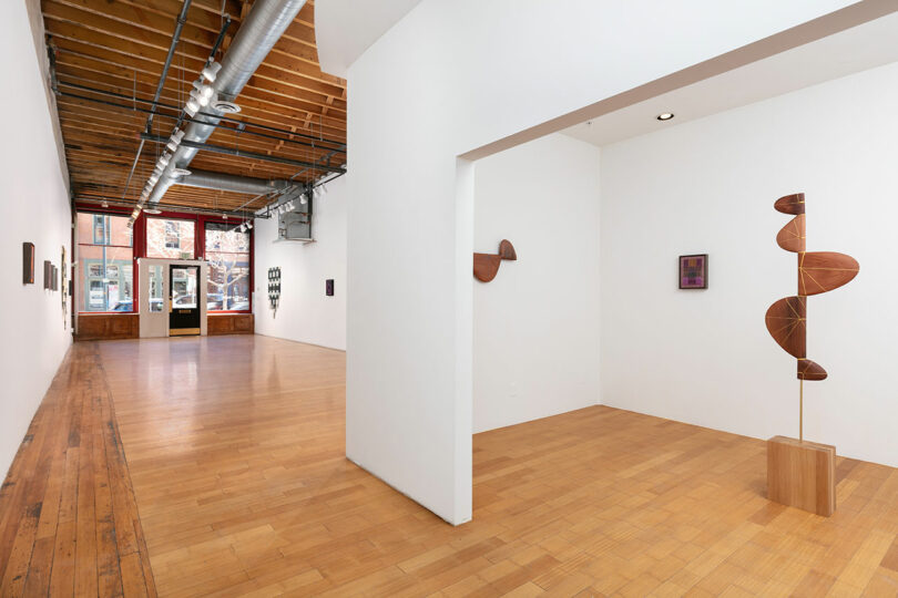 A bright art gallery interior featuring wooden floors, white walls, and modern sculptures near a window overlooking a city street.