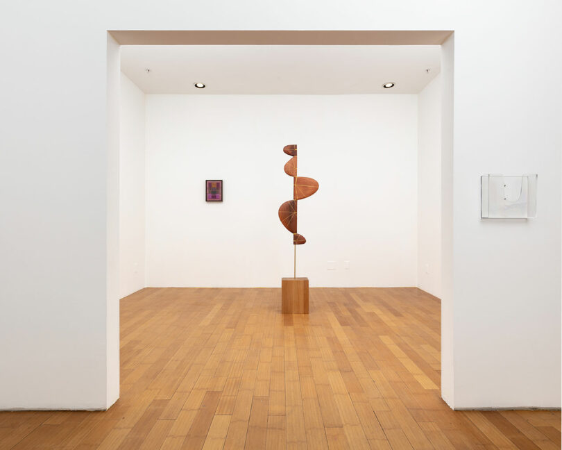 A modern art gallery interior with a large wooden spiral sculpture on a pedestal, and two framed artworks on the white walls.