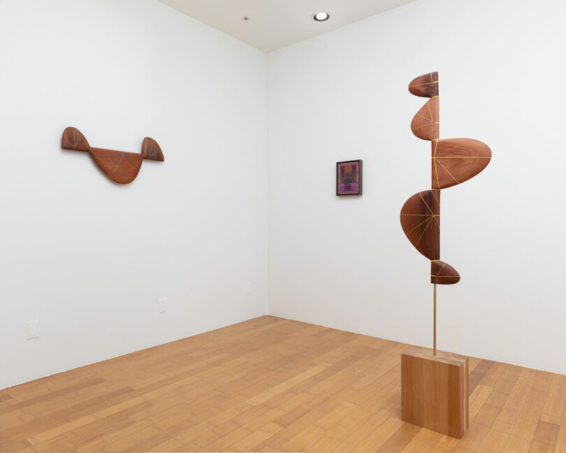 Art gallery corner with three abstract wooden sculptures: one freestanding helix-shaped piece and two wall-mounted shapes resembling wings.