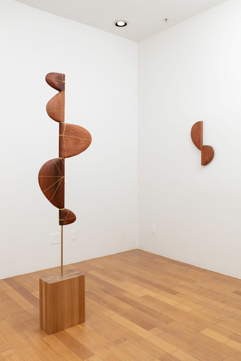 Sculpture installation in a gallery featuring a tall, vertical piece with curved wooden elements, and a smaller, round wooden wall piece.
