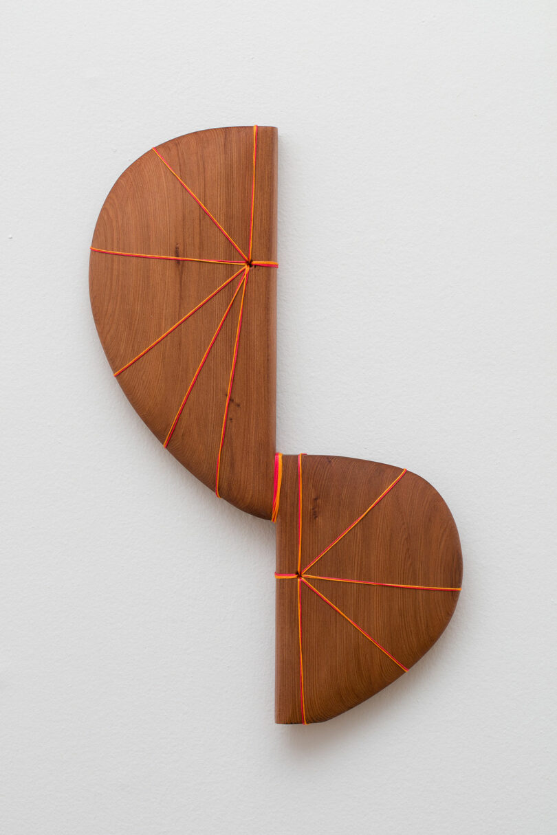 A wooden sculpture shaped like a broken heart, composed of two pieces bound together with red thread, mounted on a white wall.