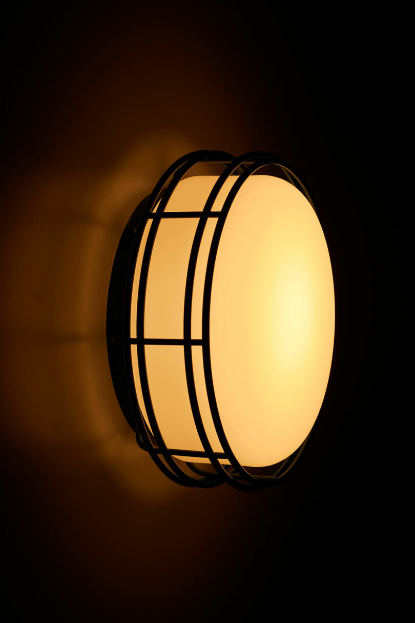 A wall-mounted round light fixture with a glowing yellow bulb, encased in a black metal frame against a dark background.