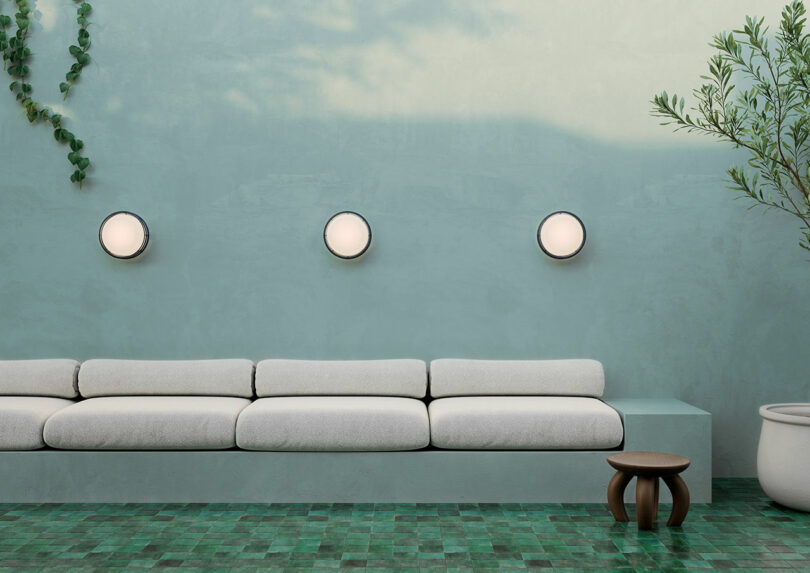 Minimalist living room with a long white sofa, round wall lights, a wooden stool, and potted plants against a pastel teal wall.