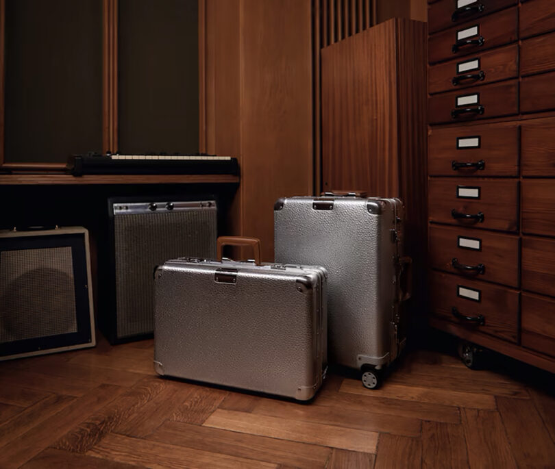 Two Rimowa metal suitcases and a briefcase in a room with wooden paneling and an amplifier.