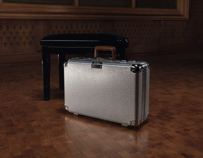 Silver Rimowa luggage briefcase on a wooden floor with a black grand piano in the background.