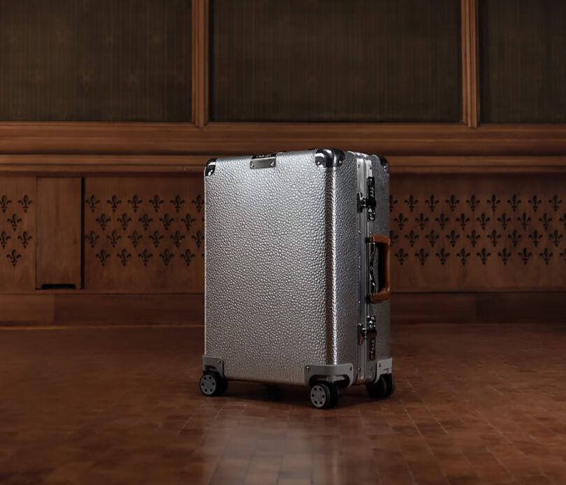 A silver Rimowa Cabin suitcase standing upright on a wooden floor against a paneled wall.