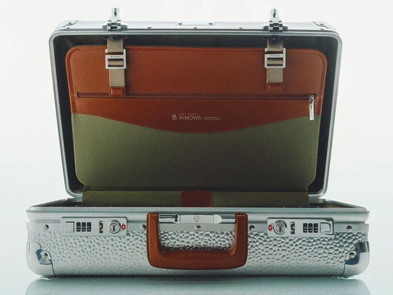Open Rimowa hammered texture aluminum suitcase with brown leather interior detailing against a light background.