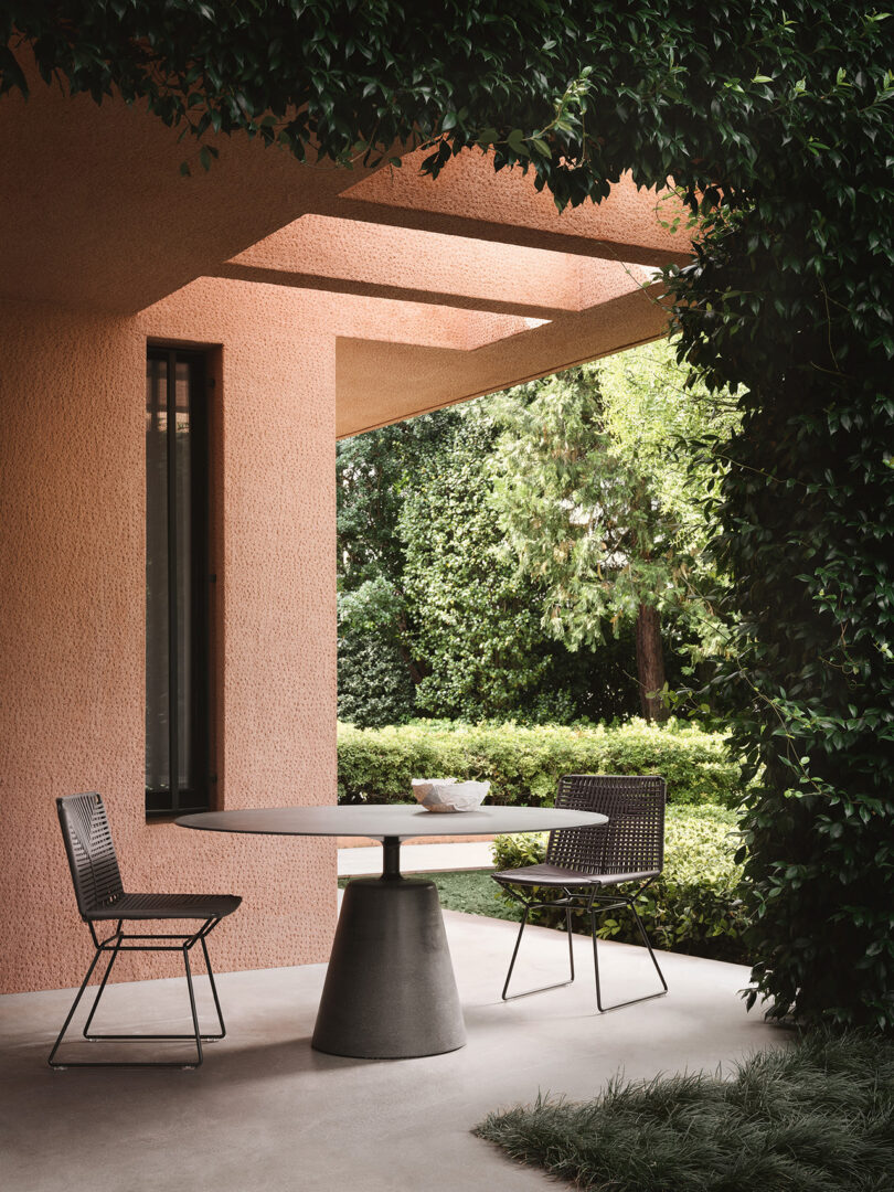 Modern outdoor seating area with two chairs and a table under an architectural overhang of a pink textured wall, surrounded by lush greenery.