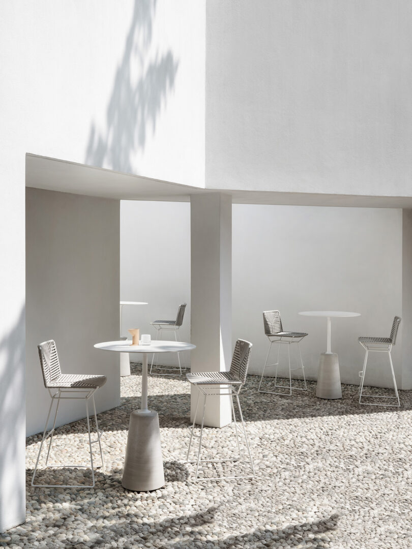 Minimalist outdoor space with white tables and chairs on gravel, shaded by white concrete walls and partial roofing, with a pattern of leaf shadows.